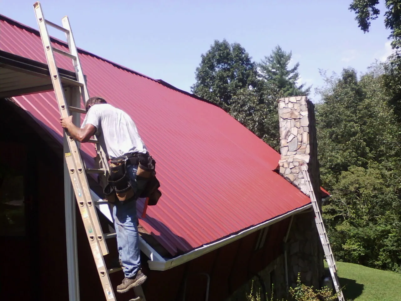 Roofer on a ladder beside a red metal roof and stone chimney — Soffit & Fascia Repair by Roofing Brothers in Benton City